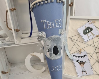 May include: A blue school cone with a gray geometric patterned top, adorned with a koala bear applique. The cone has the name "THIES" and the year "2023" embroidered on it. A decorative item for the first day of school.