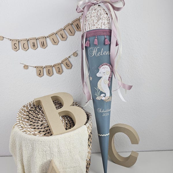 Può includere: Un cono scolastico blu con un design a cavalluccio marino, personalizzato con il nome "Helen" e l'anno "2026". Il cono è decorato con nastri e nappe e si trova vicino a lettere di legno e uno striscione.