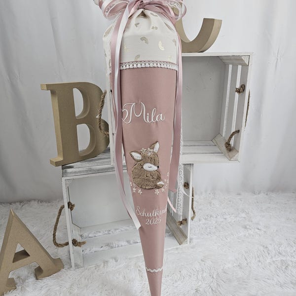 Può includere: Un cono scolastico rosa e crema con un disegno di cervo e il nome "Mila" ricamato. Il cono è decorato con nastri e pizzo, ed è visibile anche il testo "Schulkind 2023".