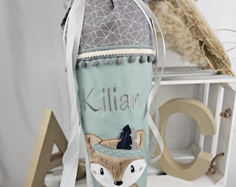 May include: A mint green cone-shaped school bag with a fox design and the name "Kilian" embroidered in grey. The bag is topped with a grey geometric patterned fabric and tied with a silver ribbon. The bag has a braided trim and pom-poms.