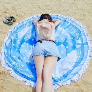 Pode incluir: Uma toalha de praia redonda com um padr&atilde;o azul e branco, bordas com franjas e uma pessoa deitada sobre ela. A toalha tem um fundo azul claro com bolinhas brancas e formas abstratas. Um chap&eacute;u de palha repousa nas costas da pessoa.