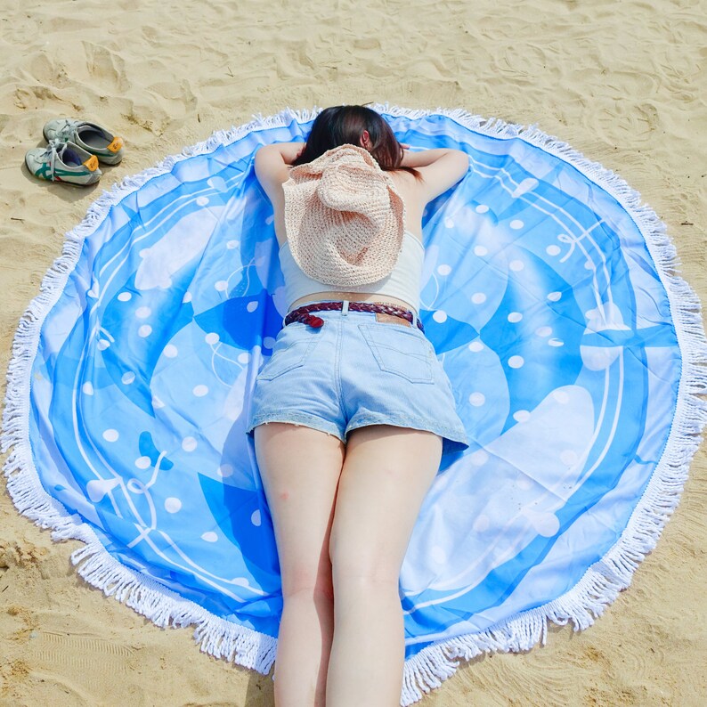 Pode incluir: Uma toalha de praia redonda com um padr&atilde;o azul e branco, bordas com franjas e uma pessoa deitada sobre ela. A toalha tem um fundo azul claro com bolinhas brancas e formas abstratas. Um chap&eacute;u de palha repousa nas costas da pessoa.
