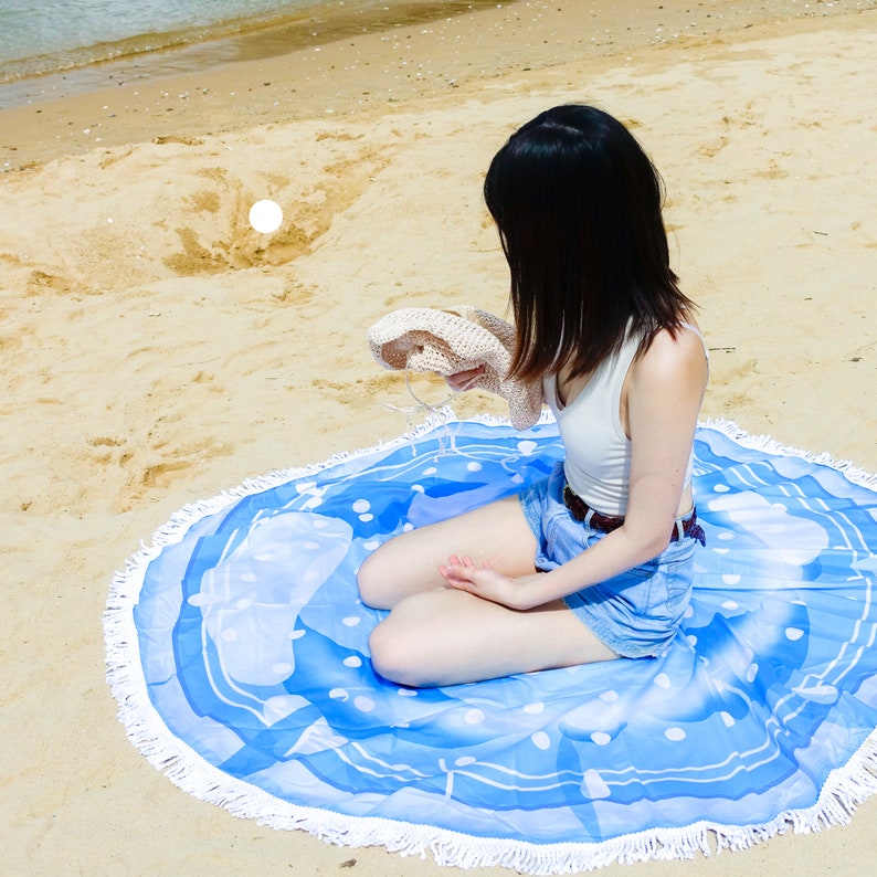 Pode incluir: Uma toalha de praia redonda com um padr&atilde;o azul e branco e uma borda de franjas brancas. A toalha est&aacute; em uma praia arenosa. Uma pessoa est&aacute; sentada na toalha, segurando um chap&eacute;u de sol bege.