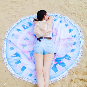 Puede incluir: Una toalla de playa redonda con borde de flecos blancos. La toalla presenta un dise&ntilde;o azul y rosa sobre un fondo blanco. Un sombrero de paja descansa sobre la toalla.