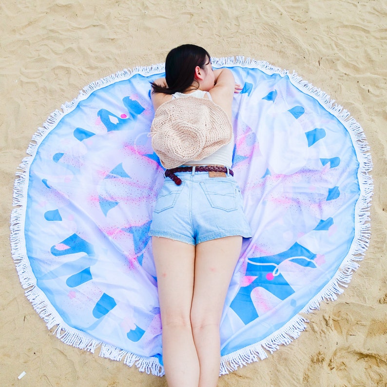 Puede incluir: Una toalla de playa redonda con borde de flecos blancos. La toalla presenta un dise&ntilde;o azul y rosa sobre un fondo blanco. Un sombrero de paja descansa sobre la toalla.