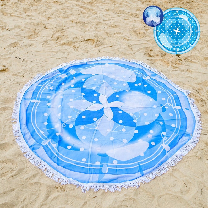 Pode incluir: Toalha de praia redonda com um design de &aacute;gua-viva azul e branco. A toalha apresenta um padr&atilde;o floral central, bolinhas brancas e uma borda de franjas brancas. A toalha est&aacute; em uma praia arenosa.
