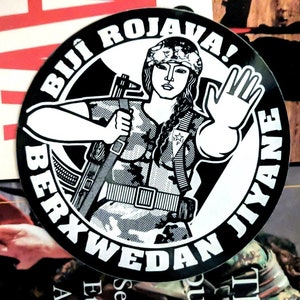 Peut inclure: Autocollant noir et blanc circulaire. Il représente une femme en tenue militaire, tenant un fusil et levant la main. Les mots "BIJI ROJAVA!" et "BERXWEDAN JIYANE" entourent l'image.