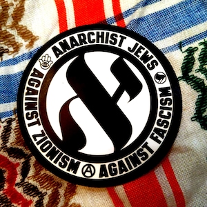 Könnte beinhalten: Schwarzer und weißer runder Aufkleber mit dem Text "Against Zionism Against Fascism Anarchist Jews" in einem Kreis um einen stilisierten Buchstaben "A".