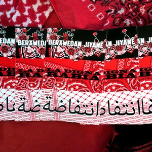 K&ouml;nnte beinhalten: Eine Sammlung roter und schwarzer Bandanas mit verschiedenen Mustern und Texten. Die Bandanas weisen florale Designs, Paisley-Prints und arabische Schrift auf. Der Text enth&auml;lt die W&ouml;rter "BERXWEDAN" und "JIYANE".