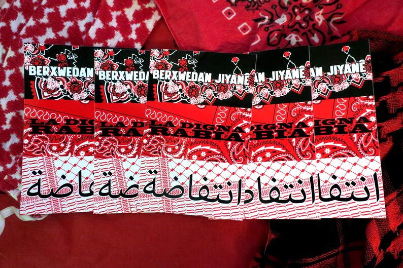K&ouml;nnte beinhalten: Eine Sammlung roter und schwarzer Bandanas mit verschiedenen Mustern und Texten. Die Bandanas weisen florale Designs, Paisley-Prints und arabische Schrift auf. Der Text enth&auml;lt die W&ouml;rter "BERXWEDAN" und "JIYANE".