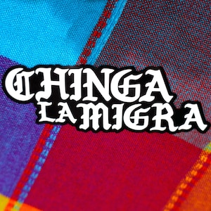 May include: A black and white sticker with the words "CHINGA LA MIGRA" in a gothic font. The sticker is set against a colorful background of blue, red, purple, and orange plaid fabric.
