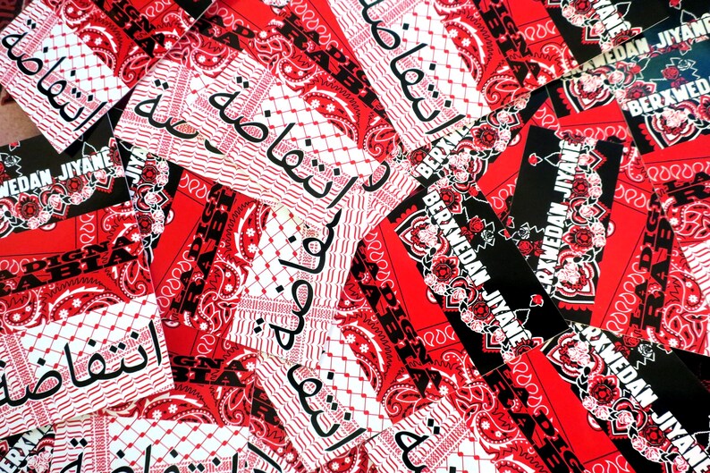K&ouml;nnte beinhalten: Eine Sammlung roter und schwarzer Bandanas und Aufkleber. Die Bandanas weisen ein Paisley-Muster und arabischen Text auf. Einige Aufkleber tragen den wei&szlig;en Text "BERXWEDAN JIYANE". Das Gesamtthema ist politischer Aktivismus.