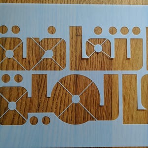 Może przedstawiać: Biała szablon z wyciętym arabskim słowem "Salam". Szablon znajduje się na drewnianej powierzchni.