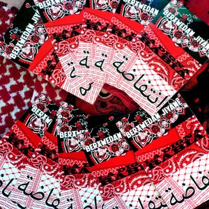 K&ouml;nnte beinhalten: Eine Sammlung roter und schwarzer Bandanas mit wei&szlig;en Paisley-Mustern und arabischem Text. Einige Bandanas tragen den wei&szlig;en Text "BERXWEDAN JIYANE". Die Bandanas sind f&auml;cherf&ouml;rmig angeordnet.