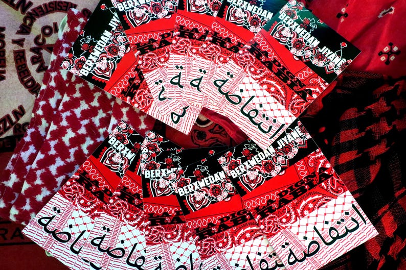 K&ouml;nnte beinhalten: Eine Sammlung roter und schwarzer Bandanas mit wei&szlig;en Paisley-Mustern und arabischem Text. Einige Bandanas tragen den wei&szlig;en Text "BERXWEDAN JIYANE". Die Bandanas sind f&auml;cherf&ouml;rmig angeordnet.