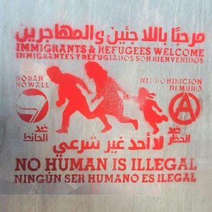 Op de afbeelding: Rode sjabloonkunst op een wit oppervlak met de tekst "Welkom immigranten en vluchtelingen" in het Engels, Spaans en Arabisch. De tekst "Geen mens is illegaal" is ook in het Engels en Spaans opgenomen. De kunst toont een gestileerde afbeelding van drie mensen die rennen, een anarchistisch symbool en een vredesteken.