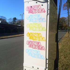 Puede incluir: Una caja de servicios p&uacute;blicos blanca con las palabras "CHINGA LA MIGRA" en varios colores, incluyendo rosa, azul, amarillo y rojo. La caja est&aacute; montada en un poste de madera, con un cielo azul de fondo.