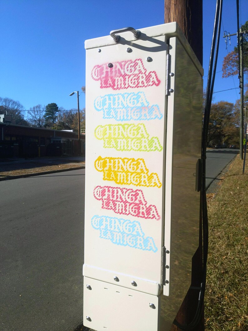 Puede incluir: Una caja de servicios p&uacute;blicos blanca con las palabras "CHINGA LA MIGRA" en varios colores, incluyendo rosa, azul, amarillo y rojo. La caja est&aacute; montada en un poste de madera, con un cielo azul de fondo.