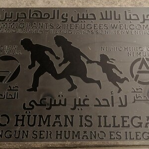 Op de afbeelding: Een zilveren bord met zwarte tekst en afbeeldingen. Het bord luidt "Welkom immigranten en vluchtelingen" in het Engels en Spaans. Het zegt ook "Geen mens is illegaal" in het Engels en Spaans. Het bord toont een afbeelding van een familie die loopt, een wereldbol met de woorden "No Wall" en een anarchistisch symbool.