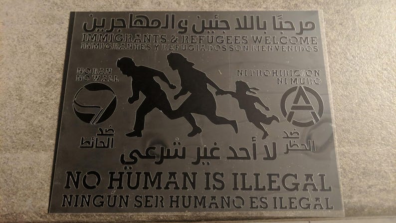 Op de afbeelding: Een zilveren bord met zwarte tekst en afbeeldingen. Het bord luidt "Welkom immigranten en vluchtelingen" in het Engels en Spaans. Het zegt ook "Geen mens is illegaal" in het Engels en Spaans. Het bord toont een afbeelding van een familie die loopt, een wereldbol met de woorden "No Wall" en een anarchistisch symbool.