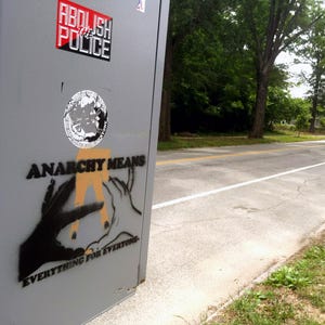 May include: A gray metal box with stickers and graffiti. The stickers read "ABOLISH THE POLICE" and feature a circular design. Graffiti on the box says "ANARCHY MEANS EVERYTHING FOR EVERYONE" with hand illustrations.