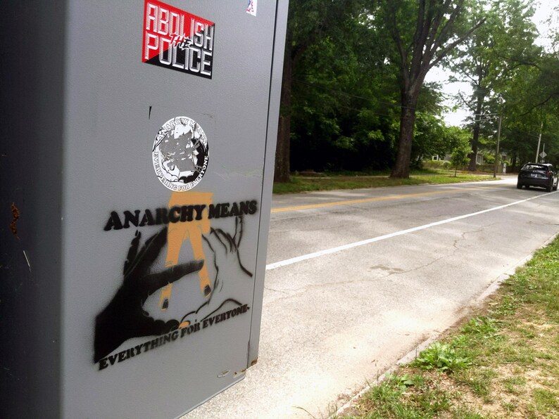 May include: A gray metal box with stickers and graffiti. The stickers read "ABOLISH THE POLICE" and feature a circular design. Graffiti on the box says "ANARCHY MEANS EVERYTHING FOR EVERYONE" with hand illustrations.