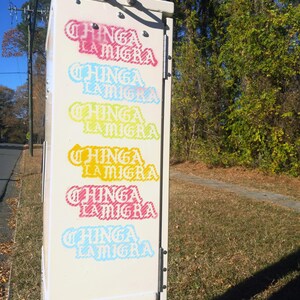 Puede incluir: Una caja de utilidad beige con las palabras "CHINGA LA MIGRA" en varios colores, incluyendo rosa, azul y amarillo. La caja est&aacute; al aire libre, con una carretera y &aacute;rboles en el fondo.