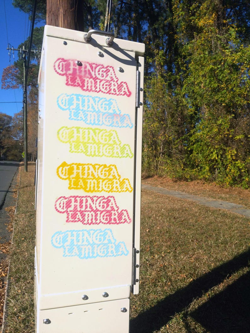 Puede incluir: Una caja de utilidad beige con las palabras "CHINGA LA MIGRA" en varios colores, incluyendo rosa, azul y amarillo. La caja est&aacute; al aire libre, con una carretera y &aacute;rboles en el fondo.