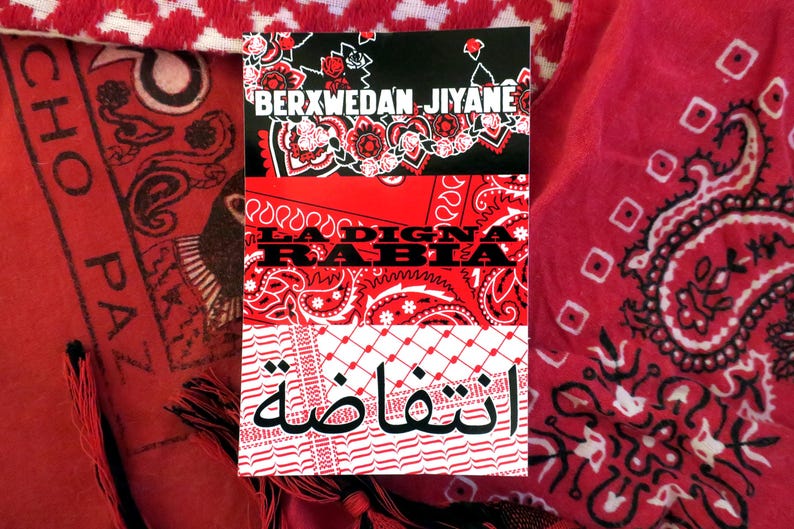 K&ouml;nnte beinhalten: Ein Stapel aus drei rot-wei&szlig;en Bandanas mit Text in Englisch und Arabisch. Das obere Bandana ist schwarz mit wei&szlig;en floralen Designs und dem Text "BERXWEDAN JIYANE". Das mittlere Bandana ist rot mit dem Text "LA DIGNA RABIA". Das untere Bandana ist wei&szlig; mit arabischem Text.