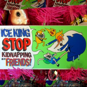 May include: A rectangular sticker with the text "ICE KING STOP KIDNAPPING OUR FRIENDS!" in blue, red, and black. The sticker features cartoon characters on a green background. The sticker is surrounded by pink tinsel.
