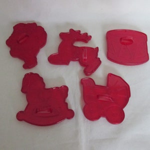 May include: Five red plastic cookie cutters in various shapes: a Santa Claus, a reindeer, a drum, a rocking horse, and a baby carriage. These cutters are ideal for making festive cookies.
