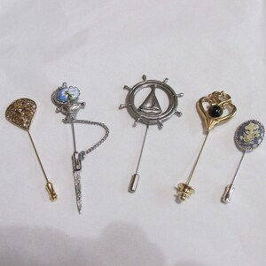 May include: A collection of five stick pins featuring diverse designs: a gold-tone filigree fan, a silver-tone floral motif with a chain, a silver-tone ship wheel with a sailboat, a gold-tone heart with a black stone, and a blue oval floral cameo.