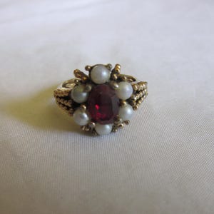 May include: A gold-toned ring featuring a large, faceted, oval-shaped red gemstone at the center, surrounded by small, round, white pearls. The ring's band has a braided design.