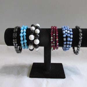 May include: Six bracelets displayed on a black T-bar stand. The collection includes a dark metallic square-bead bracelet, a light blue bead bracelet, a black and white round-bead bracelet, a red faceted-bead bracelet, a light blue bead bracelet, and a dark metallic irregular-bead bracelet.