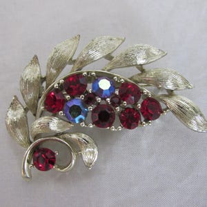 May include: Silver-toned brooch with a leaf design. The brooch features a cluster of red and blue-violet rhinestones. The leaves have a textured finish, adding detail to the vintage jewelry piece.