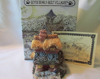 May include: A miniature cottage figurine with a thatched roof, stone walls, and a chimney. The sign reads "TED E. BEAR SHOP". The box behind the figurine has the text "BOYDS BEARLY-BUILT VILLAGES". A map of the village is also included.
