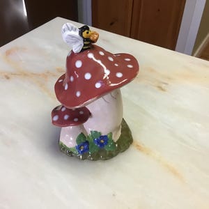 May include: A ceramic mushroom figurine with a red and white polka dot top, a white stem, and a bee on top. The mushroom is surrounded by green leaves and blue flowers.