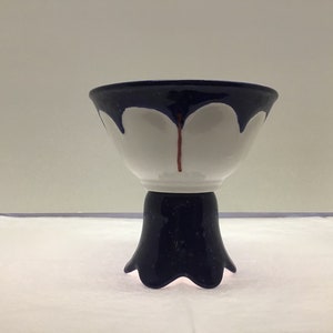 May include: A white ceramic bowl with a blue rim and a blue base. The bowl has a unique design with a blue drip effect and a red line down the center. The bowl is on a stand with a blue base and a scalloped edge.