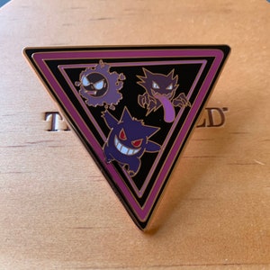 Può includere: Uno spillo smaltato triangolare con sfondo nero e bordo rosa. Lo spillo presenta tre Pokémon fantasma viola e neri: Gengar, Haunter e Gastly.