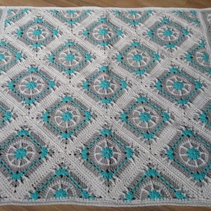 Könnte beinhalten: Eine weiße und graue Häkeldecke mit einem türkisfarbenen Rautenmuster. Die Decke hat einen Rand mit einem sich wiederholenden Blumendesign.