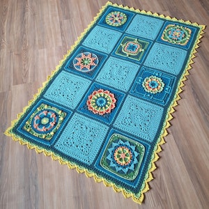 Puede incluir: Una manta de ganchillo con fondo azul y borde amarillo. La manta presenta un patrón repetitivo de motivos florales coloridos.