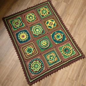 Op de afbeelding: Een vierkant gebreid deken met een bruine rand en een herhalend patroon van groene, gele en bruine bloemenmotieven.