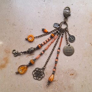 Könnte beinhalten: Ein Glücksarmband mit mehreren Anhängern, darunter eine Blume, ein Buddha, ein Friedenszeichen, ein Symbol des Lebensbaums und ein Anhänger mit der Aufschrift "live laugh love". Das Armband ist aus bronzefarbenem Metall gefertigt und hat orangefarbene und pfirsichfarbene Perlen.