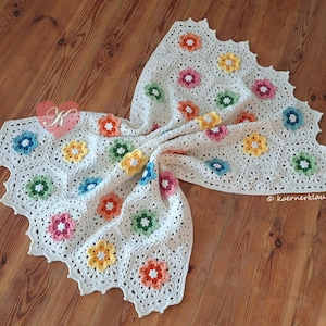 Könnte beinhalten: Eine cremefarbene Häkeldecke aus sechseckigen Motiven, die jeweils eine zentrale Blume in Orange, Gelb, Rosa, Grün und Blau zeigen. Die Decke hat einen gewellten Rand und enthält ein rotes Herz-Logo mit einem K sowie den Schriftzug koernerklaus.