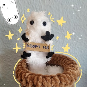 Puede incluir: Una pequeña criatura de peluche blanca y difusa con ojos y extremidades negras, que sostiene un letrero de cartón rectangular con la inscripción ADOPT ME en tinta azul. La criatura está colocada dentro de un recipiente marrón tejido.