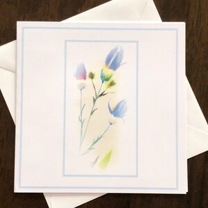 May include: White greeting card with a watercolor floral design featuring blue and purple bell-shaped flowers. The card has a light blue border and is placed on top of a white envelope, perfect for sending a thoughtful message.