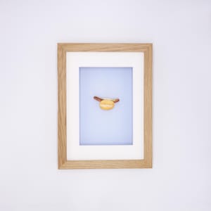 Op de afbeelding: Een ingelijste kunstdruk met een minimalistische afbeelding van een hotdog in een broodje tegen een lichtblauwe achtergrond. De hotdog is bruin en het broodje is geel. De lijst is van lichtgekleurd hout.