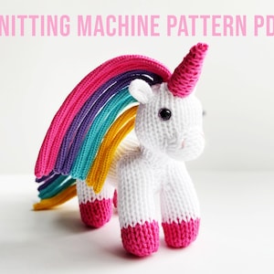 Pode incluir: Um unicórnio de tricô branco com uma juba arco-íris e cascos rosa. O unicórnio está em pé sobre uma superfície branca. Padrão de máquina de tricô PDF.