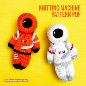 May include: Two knitted astronaut dolls, one orange and one white, with black eyes and a gray space helmet. The dolls are on a yellow background with the text "Knitting Machine Pattern PDF" and "@dianalevineknits".