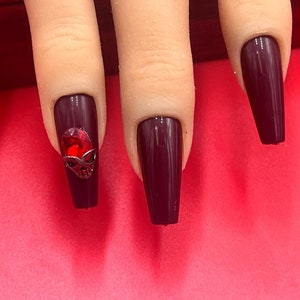 May include: Burgundy coffin-shaped press-on nails with a red rhinestone skull accent on one nail.
